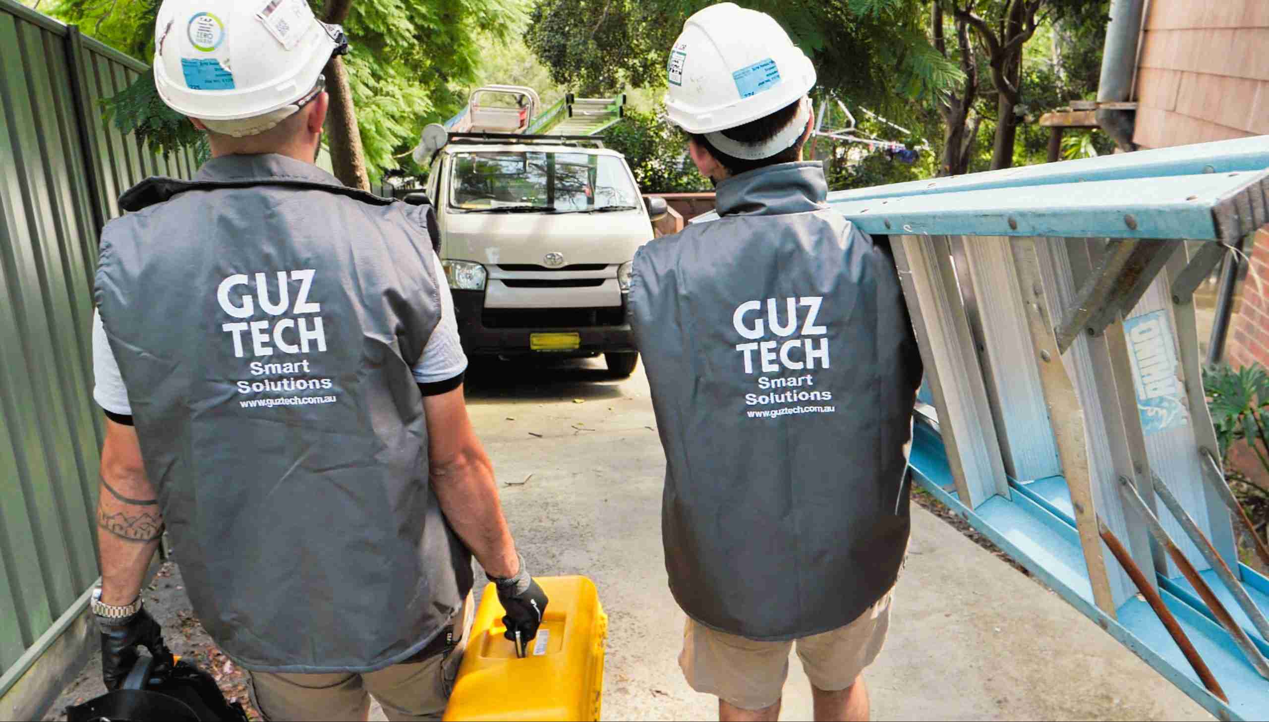 Guztech Electronic Security Technicians wearing helmets and safety gear, carrying out tools and ladders down a driveway back to their service van.